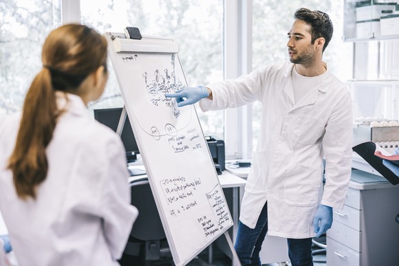 A scientist in a laboratory points to writing on a whiteboard as a coworker looks on.