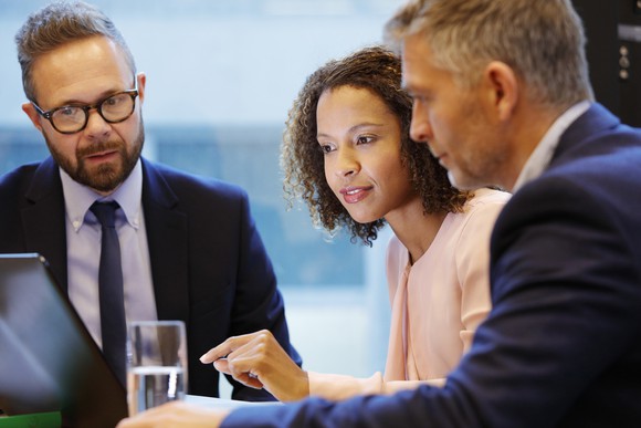 Three people in business attire are gathered around a laptop screen.