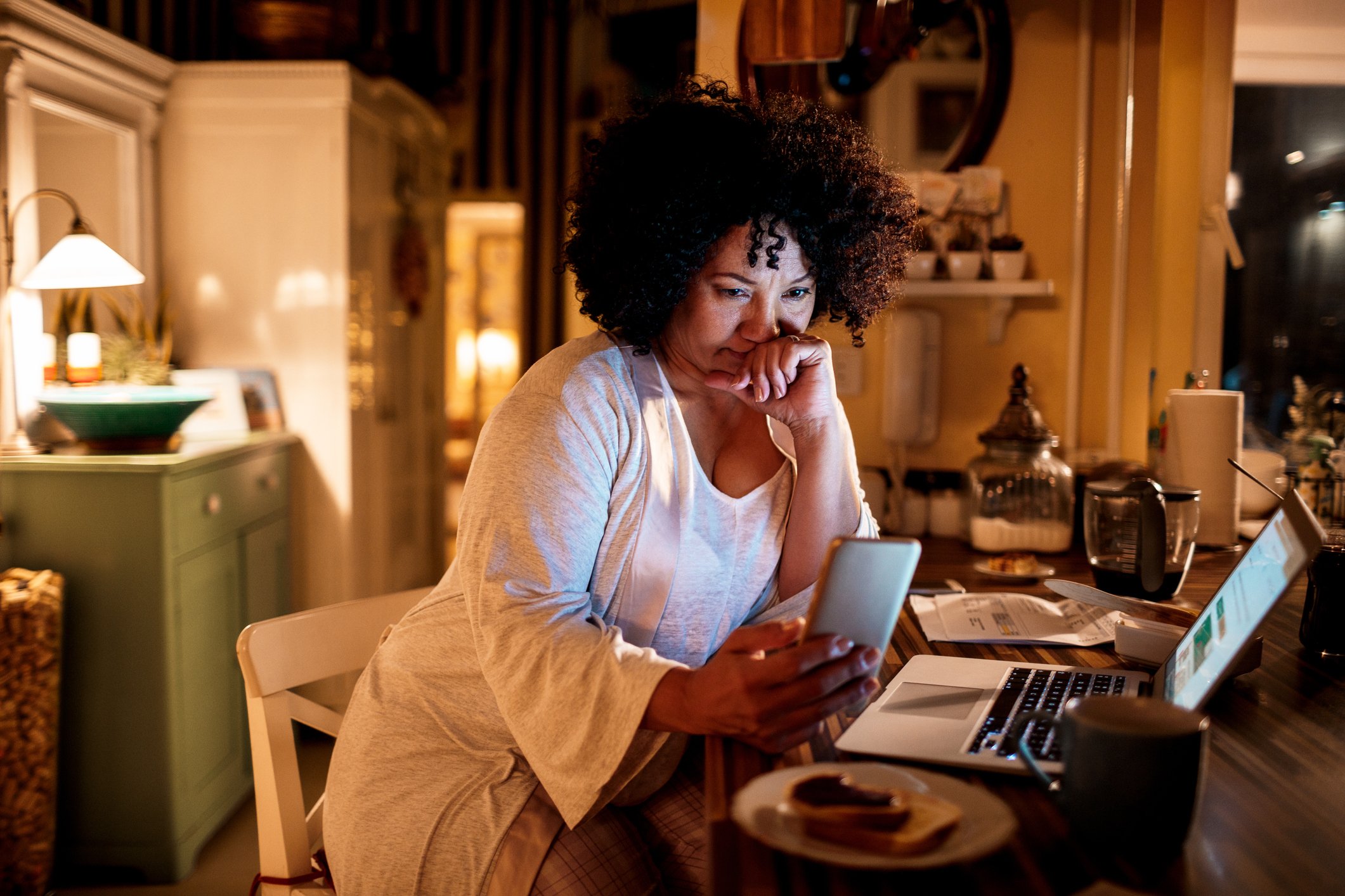 A person in their kitchen, looking at their phone, laptop on table, looking concerned.