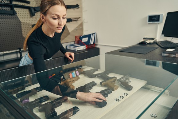 A gun store employee reaches into a glass display case full of firearms.