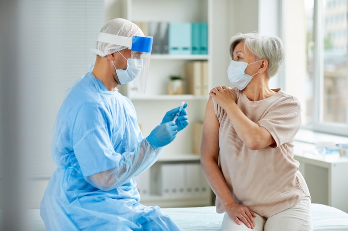 A masked, gloved, and shielded healthcare professional holding a syringe with needle and vaccine vial near the arm of a masked patient.