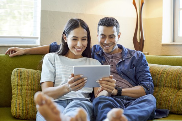 Two smiling people sitting on a sofa looking at a touchscreen tablet.