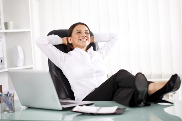 A smiling person with eyes closed leaning back in a chair with feet on desk next to a laptop.