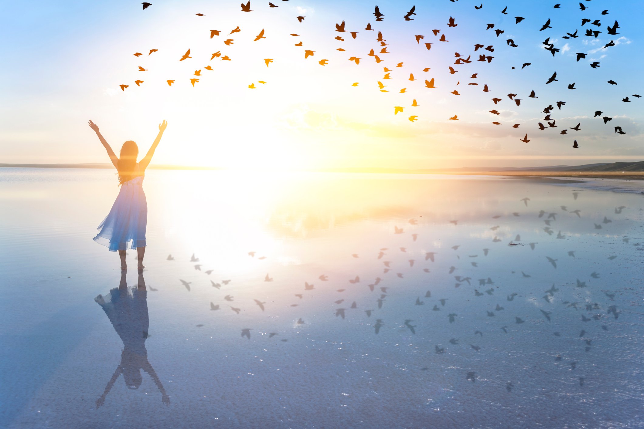 A lady lifts her arms while looking over a body of water as a flock of birds glide past during sunrise.