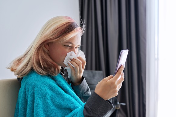 A person sitting on the sofa blowing their nose while looking at a smartphone.