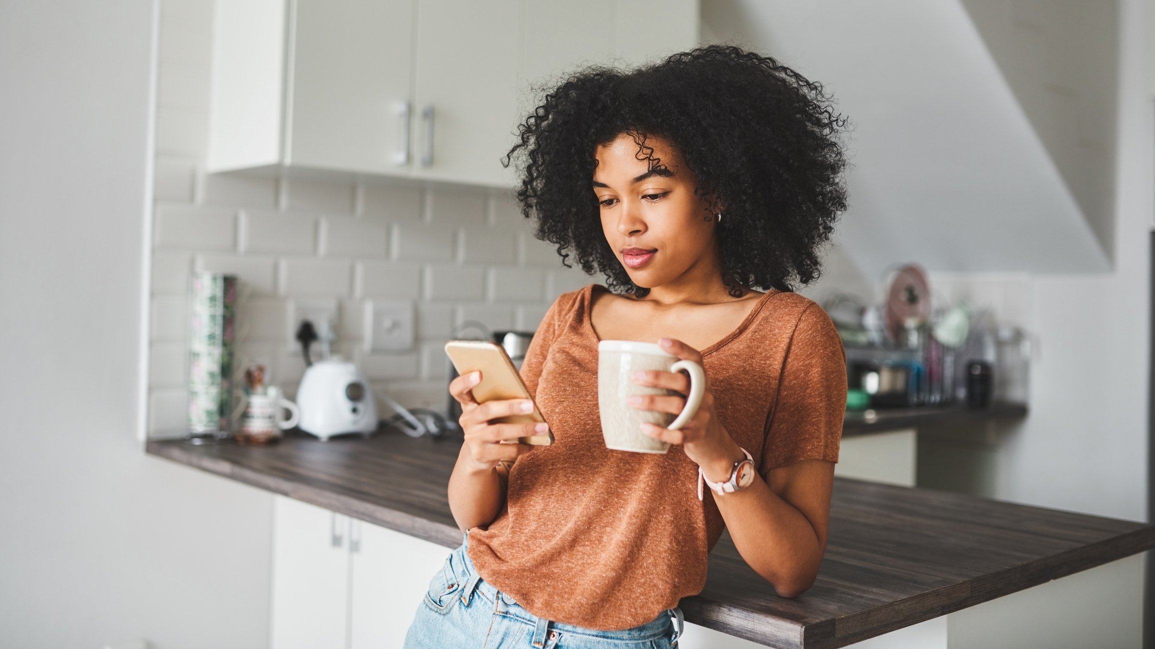 A  person looking at their phone while leaning on the kitchen counter and holding a cup of coffee in one hand.