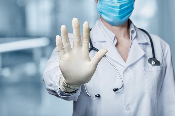 A physician wearing a face mask is holding a hand up in a halting gesture.