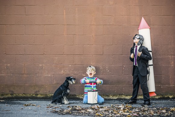 Two children play, one with rocket strapped to their back and one pretending to trigger the launch.