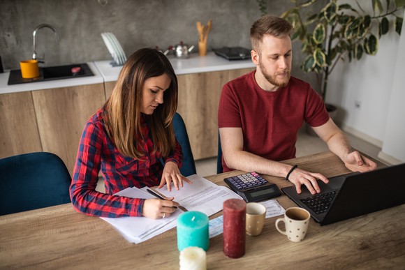 Two people at table with documents, working on laptop.