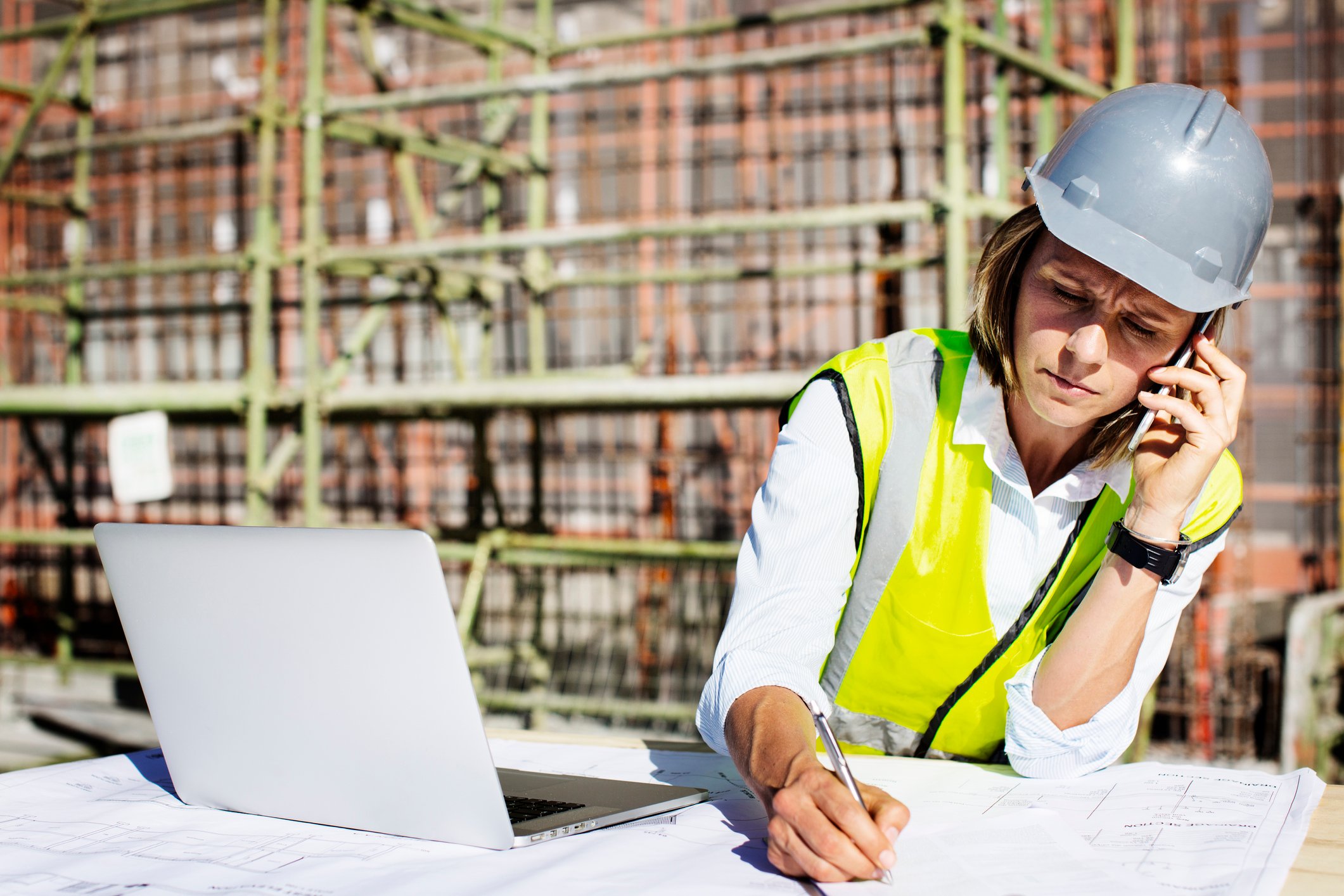 Architect on a construction site, wearing a hard hat, using a smartphone, and seated in front of a laptop.