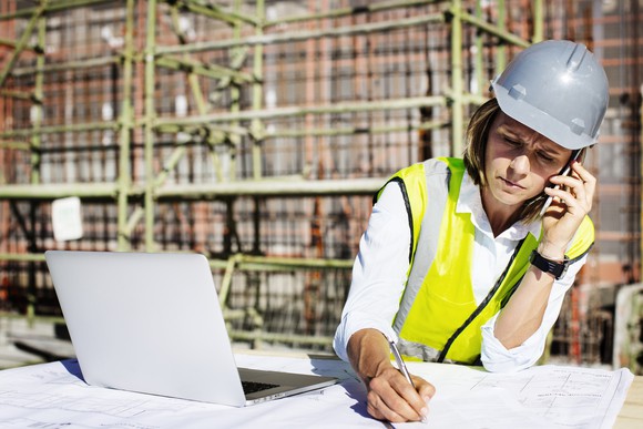 Architect on a construction site, wearing a hard hat, using a smartphone, and seated in front of a laptop.