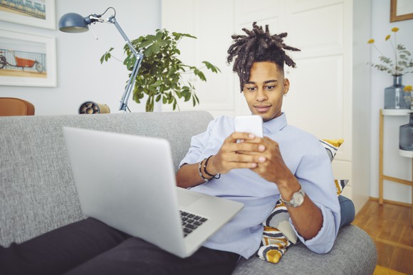 A person looks at a smartphone while balancing a laptop on a home couch.