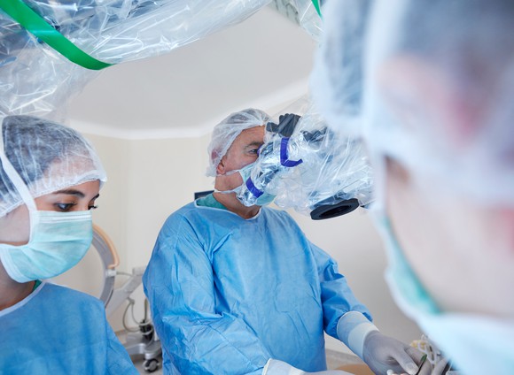Watched by a pair of doctors, a surgeon controls a robotic surgical unit as he peers into a surgical imaging system during an operation.