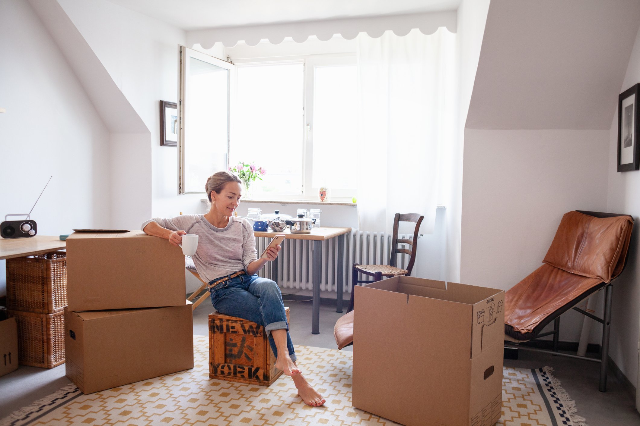 A person in an apartment sitting on a box looking at their phone.