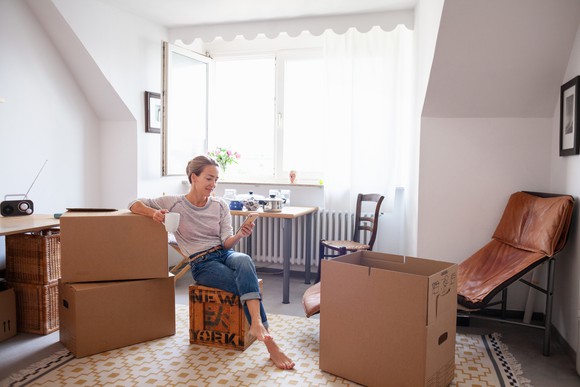 A person in an apartment sitting on a box looking at their phone.