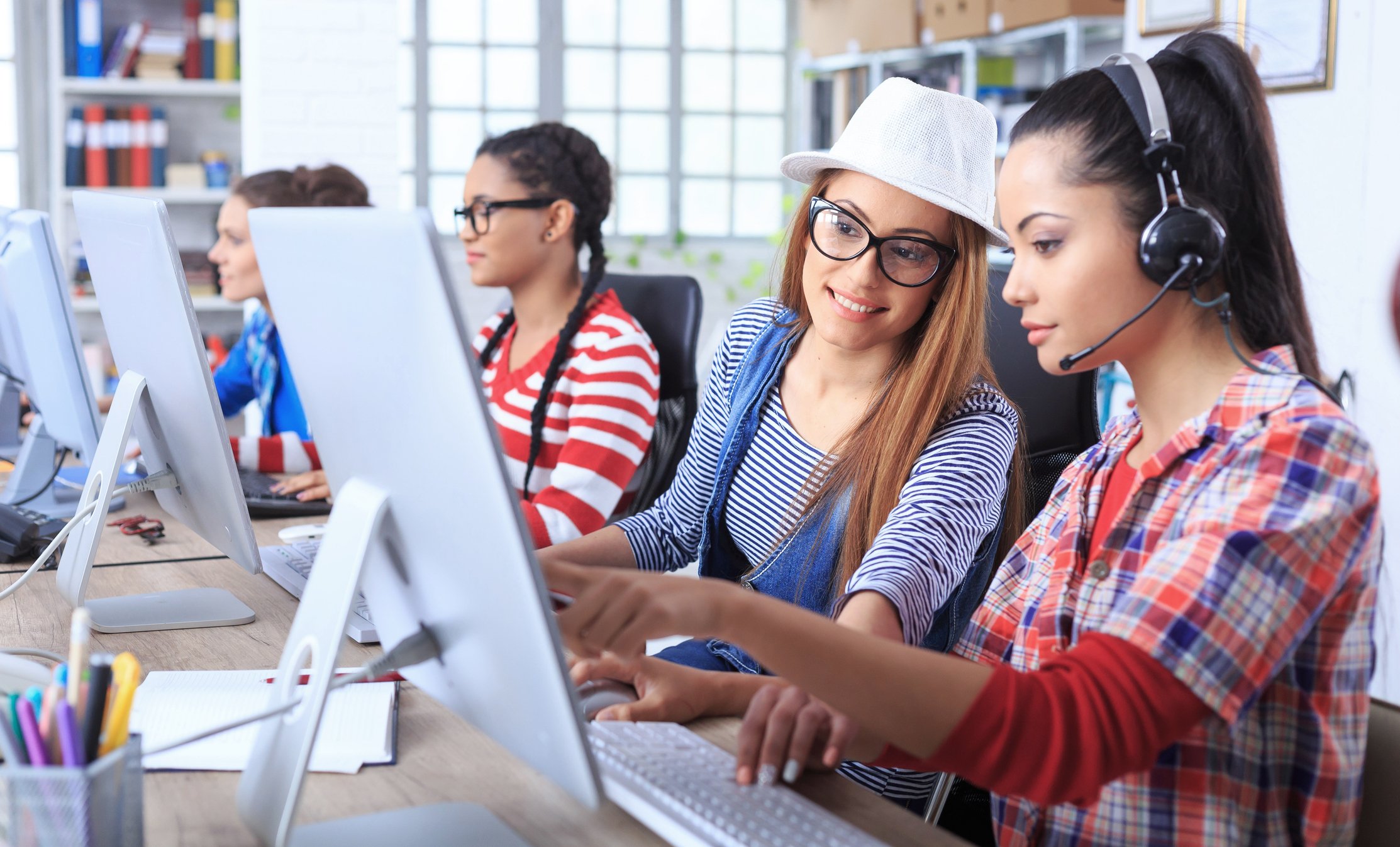 Contact center agents help each other while seated in front of monitors.
