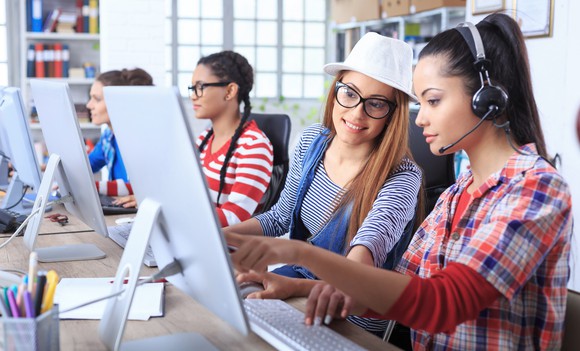 Contact center agents help each other while seated in front of monitors.