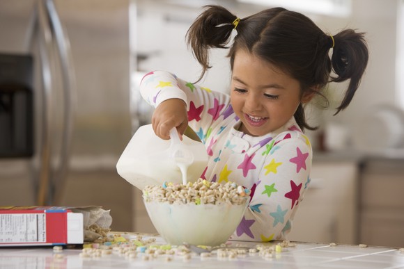 A young child pours milk into an overflowing bowl of cereal.