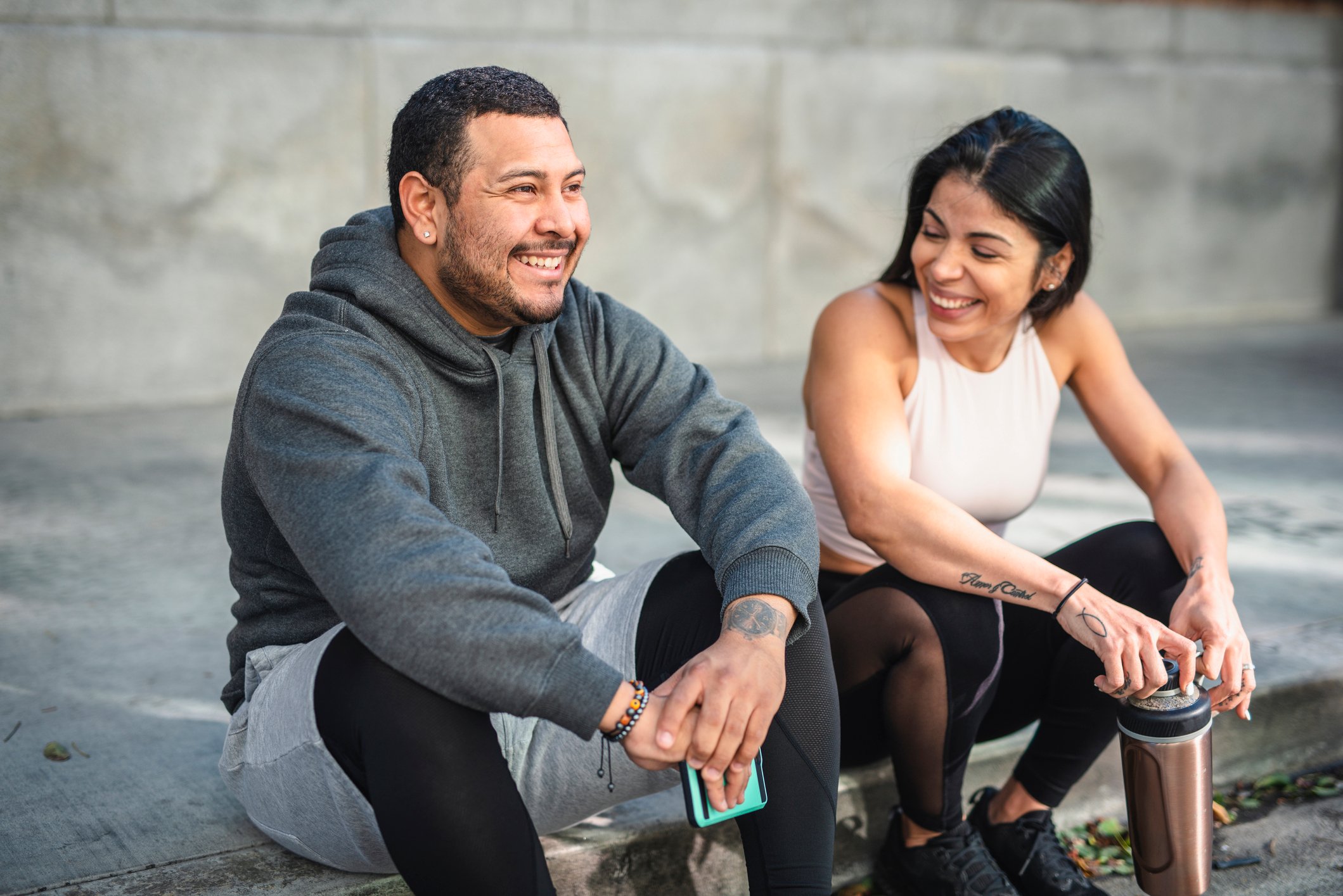 Two smiling people sitting on sidewalk wearing athletic apparel.