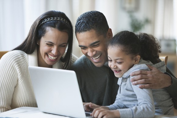 Two adults and a child smile as they look at something on a laptop in home setting.