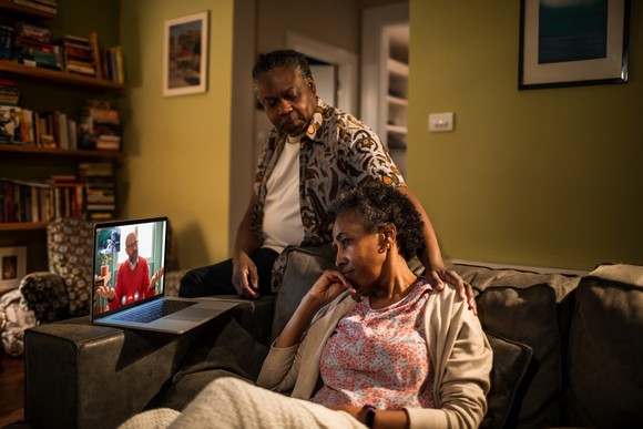 An older person sits in front of a laptop computer conducting a telehealth visit while being supported by a friend or family member.