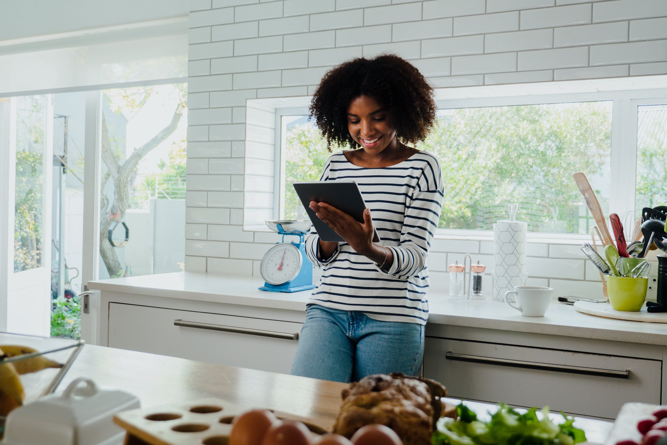 A smiling person looking at a touchscreen tablet while standing in a kitchen.