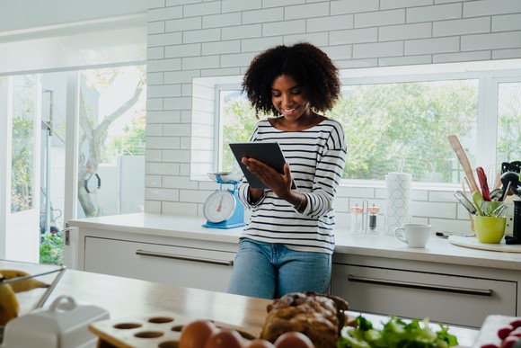A smiling person looking at a touchscreen tablet while standing in a kitchen.