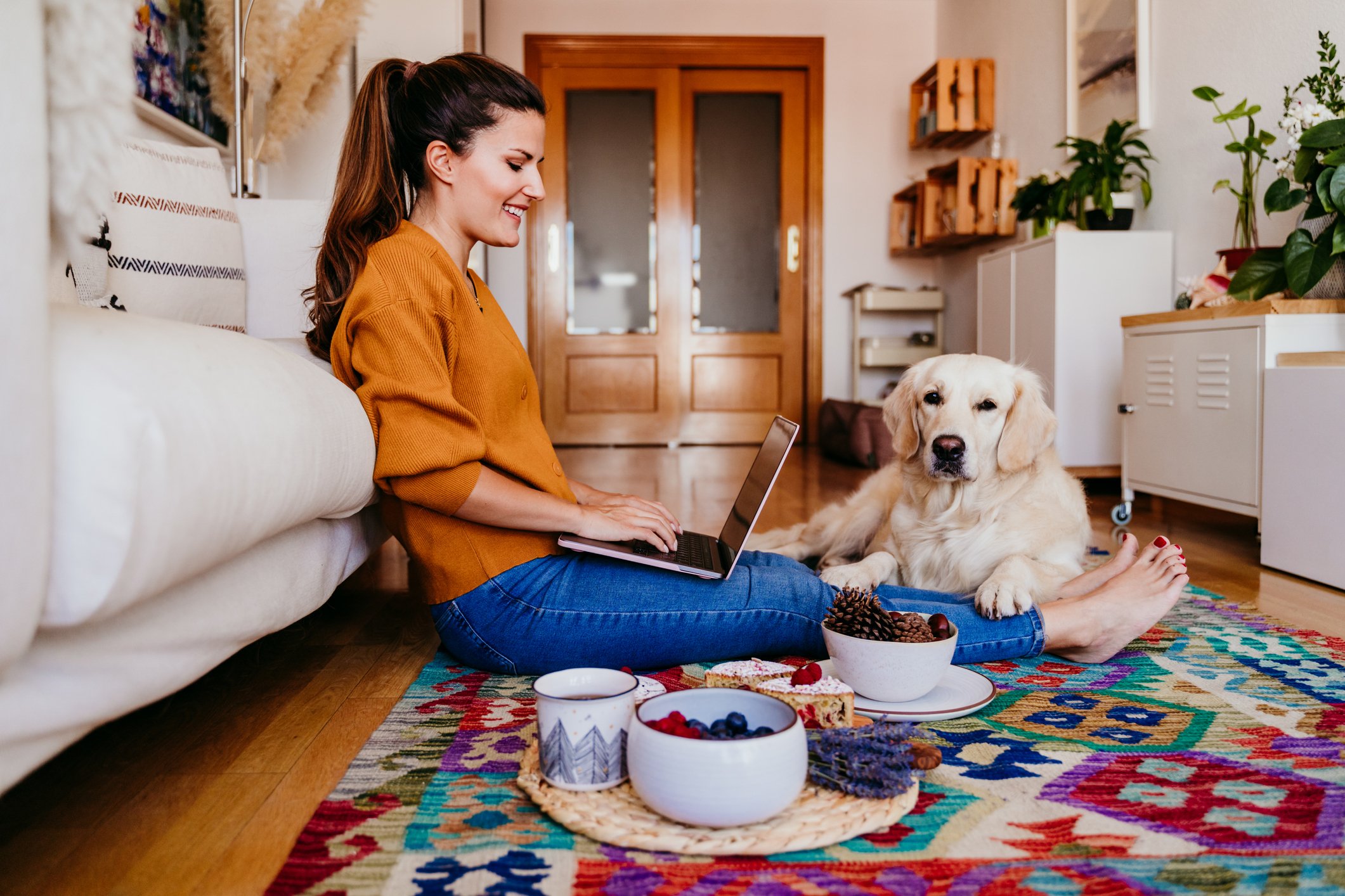 A smiling person sitting on the floor with a laptop next to a dog.