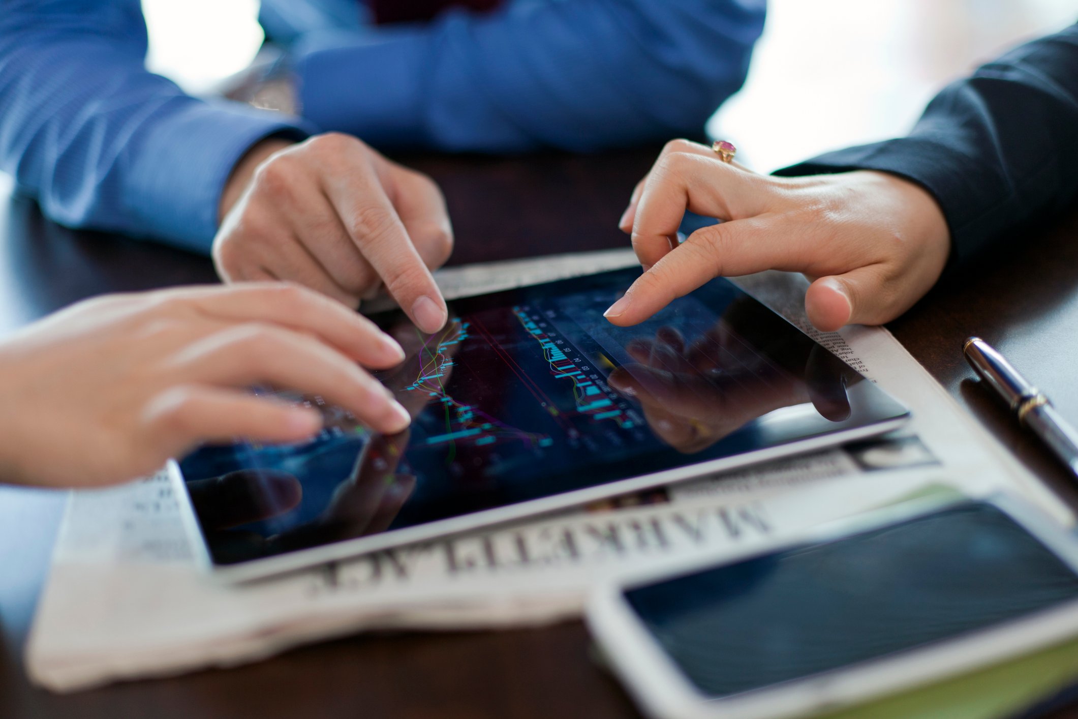 Three hands operate a tablet lying flat on a newspaper. 