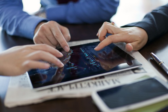 Three hands operate a tablet lying flat on a newspaper. 