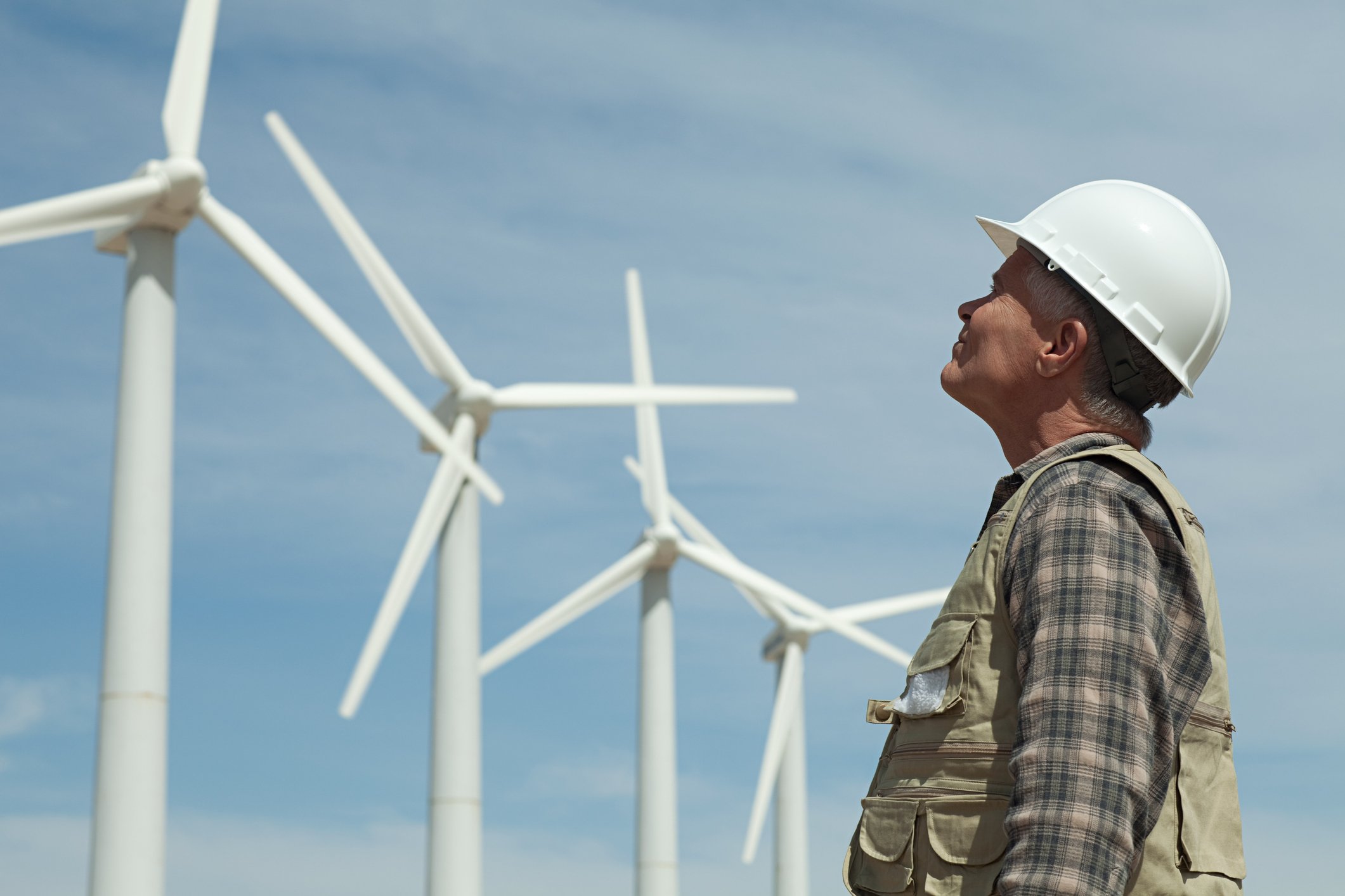 A man wearing a hard hat looks up at a row of wind turbines. 