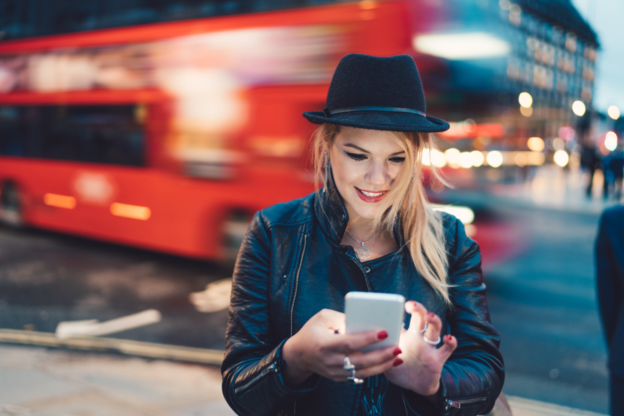 A woman wearing a hat stands on a street corner looking at a smartphone as a red bus passes behind her.