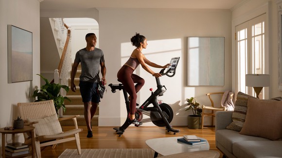 A person riding a Peloton stationary bike in a house while another person walks into the room behind them.