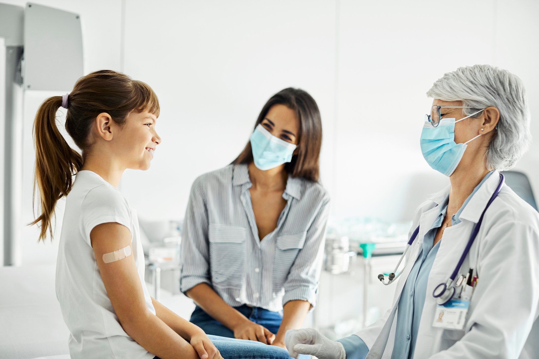 A newly vaccinated child sits on the exam table in a doctor's office facing the doctor and another adult.