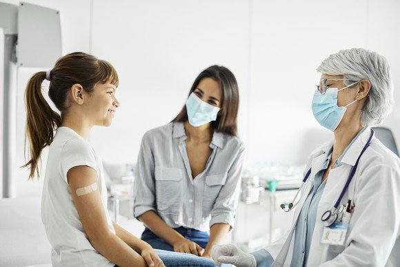 A newly vaccinated child sits on the exam table in a doctor's office facing the doctor and another adult.