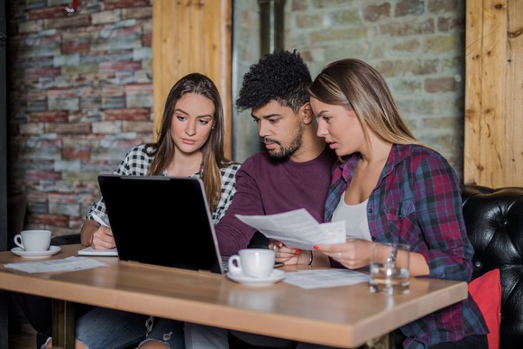 Three people sit at a table with a laptop and review papers and the screen.
