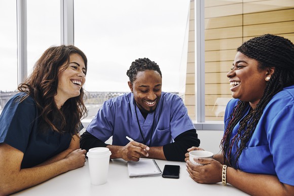 Three nurses laugh as they chat at a table.