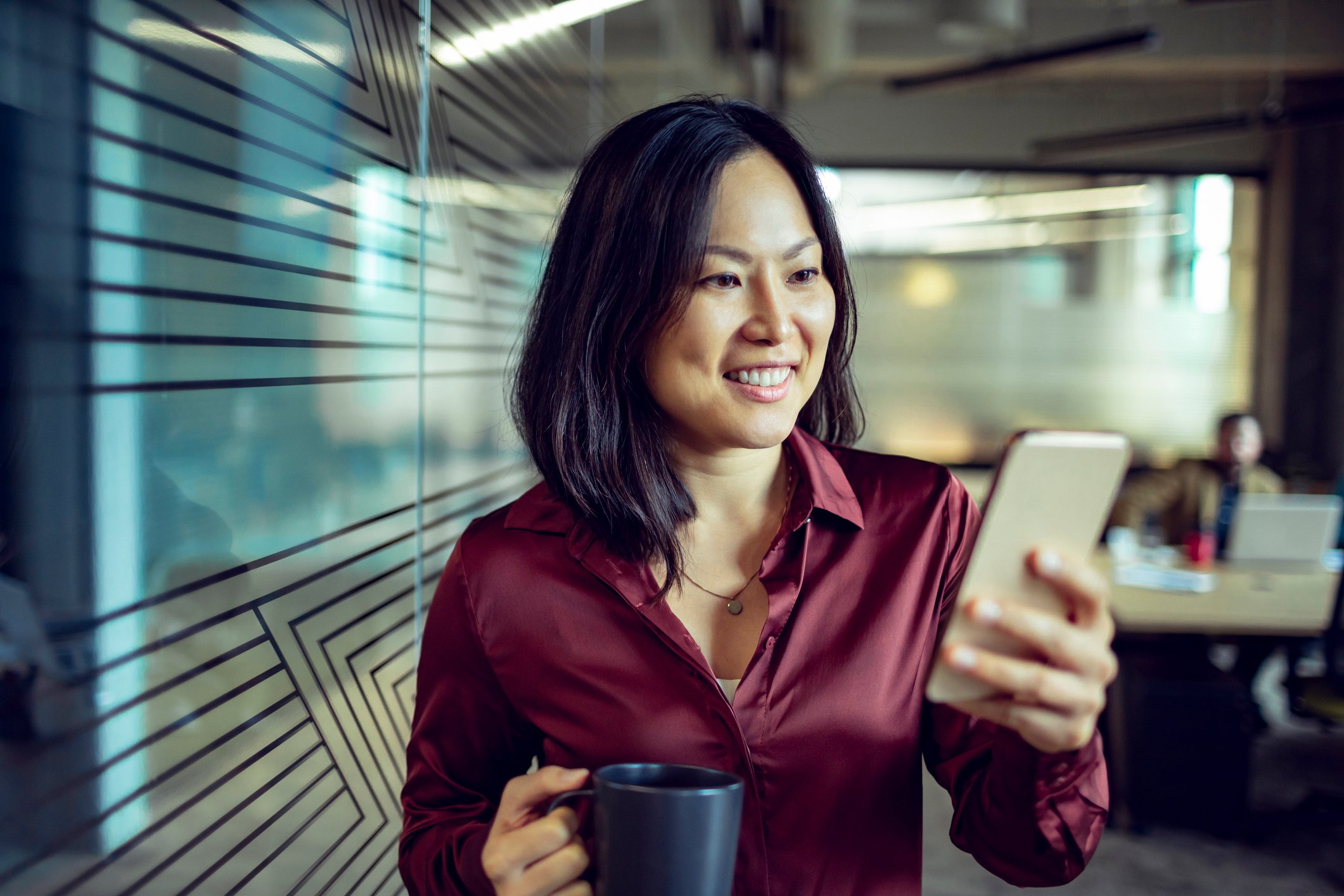 A businessperson looks at their phone while holding a cup of coffee in an office.