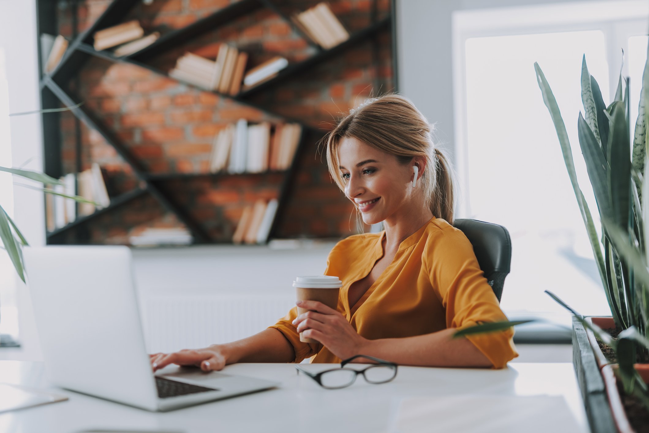 A smiling person holding a cup of coffee while looking at a laptop.