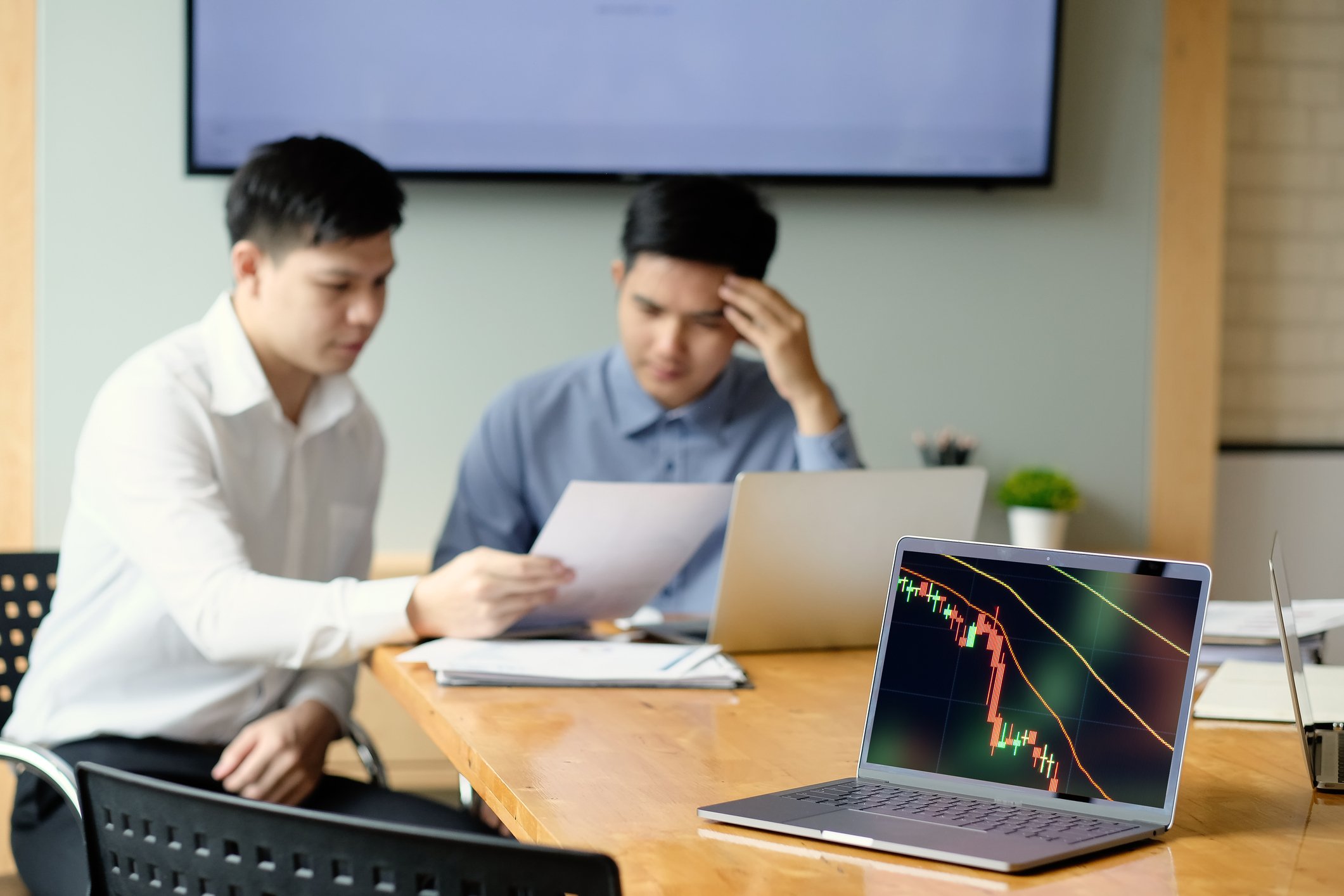 A laptop showing a stock chart trending down with two people looking at a document in the background.
