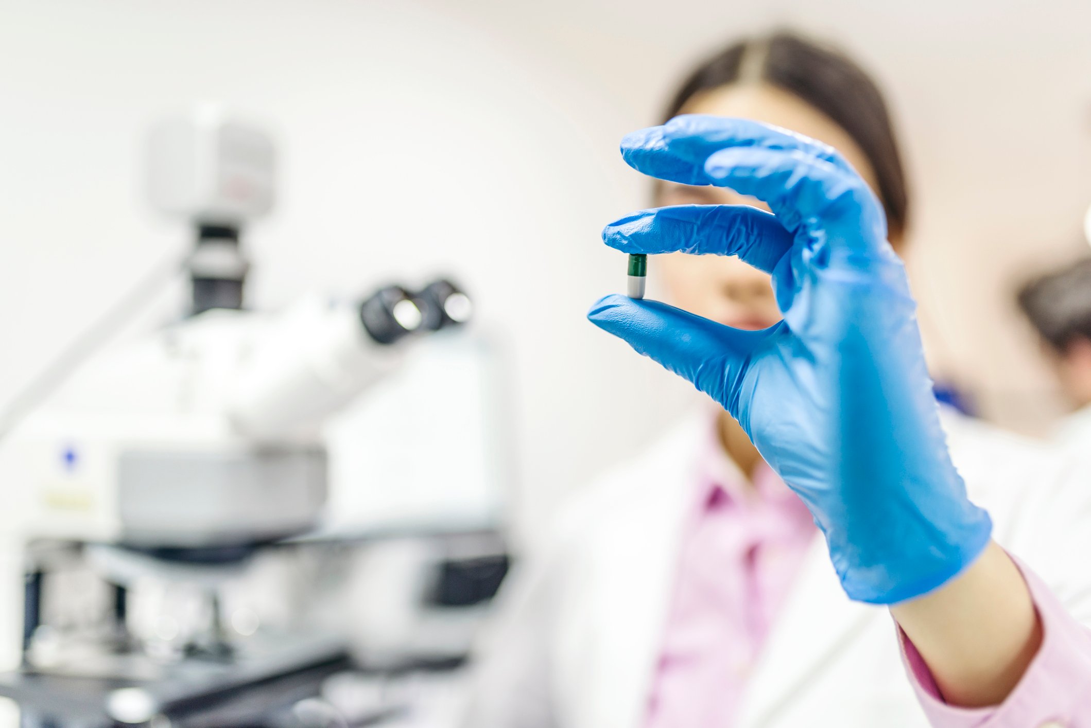 A scientist holding a pill with a microscope in the background.