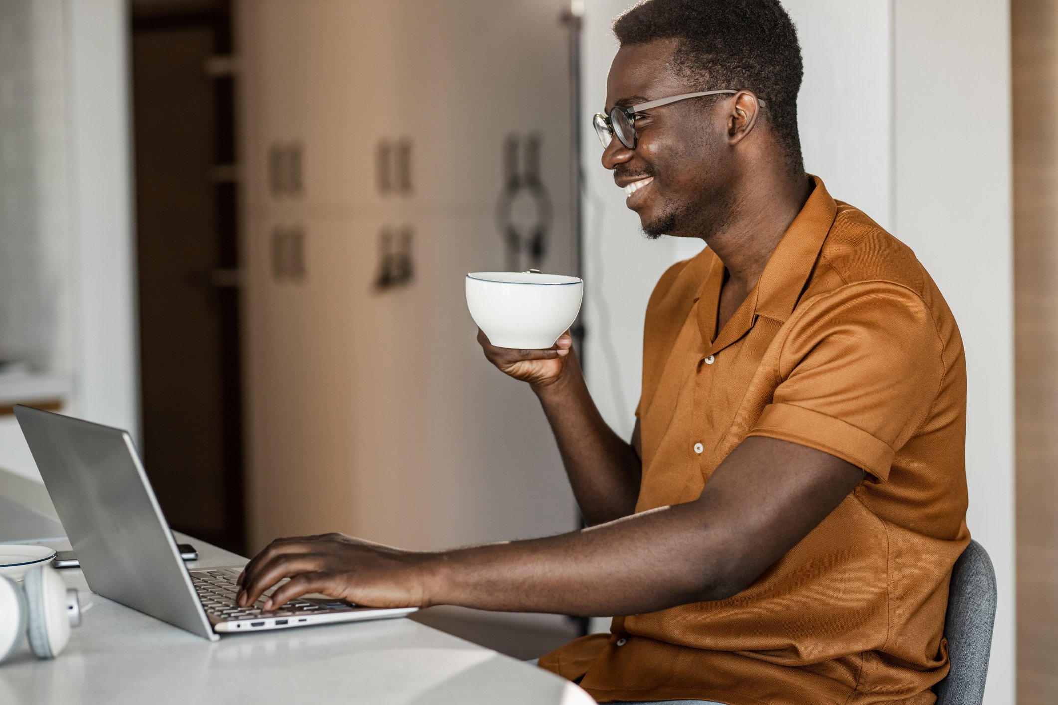 A smiling person holding a cup of coffee while looking at a laptop.
