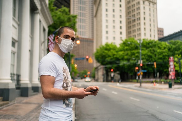 Person wearing a mask, holding a phone, and standing on the side of the road. 