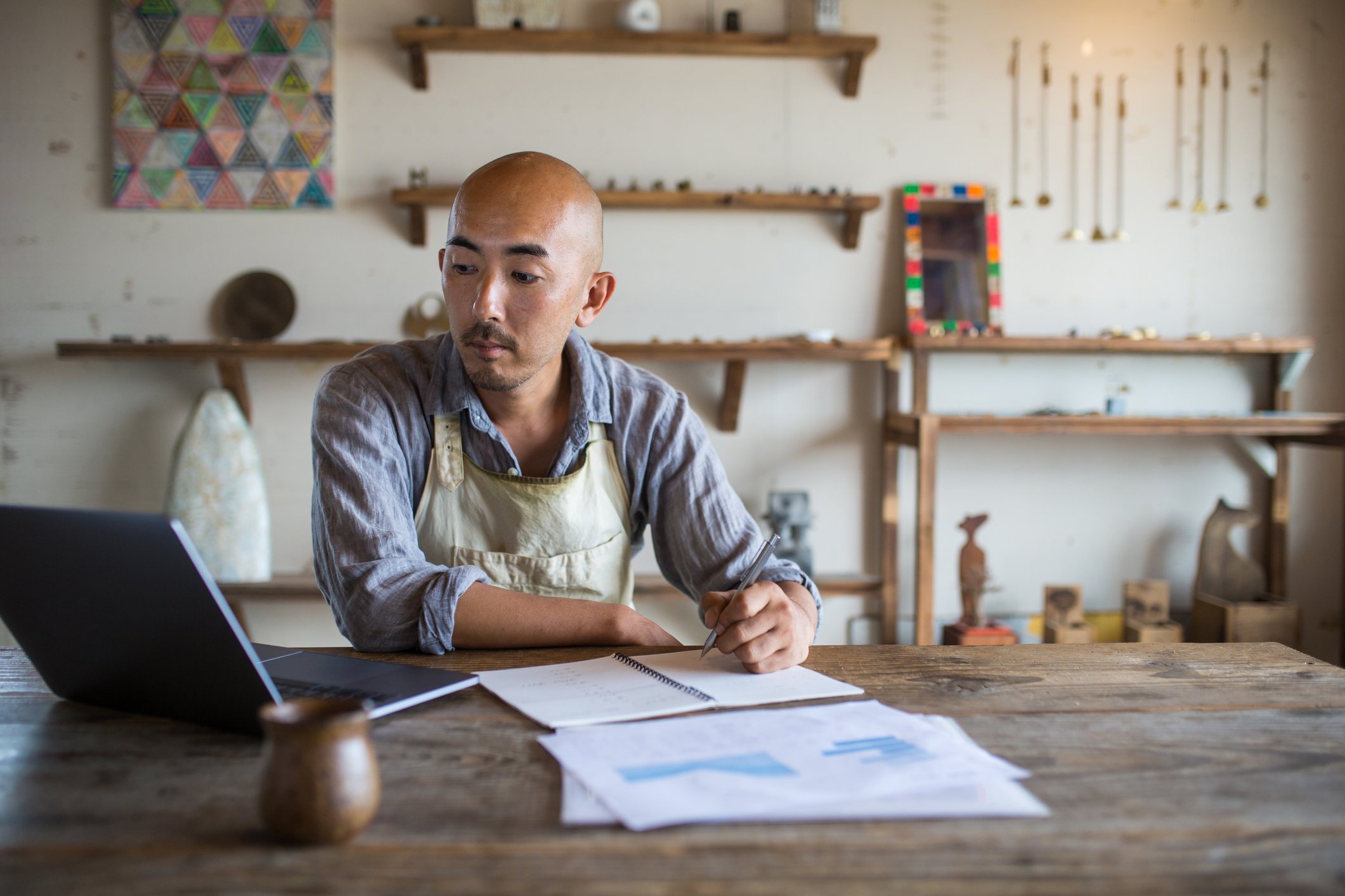 A person looks at a laptop while writing in a notebook and considering several printed charts.