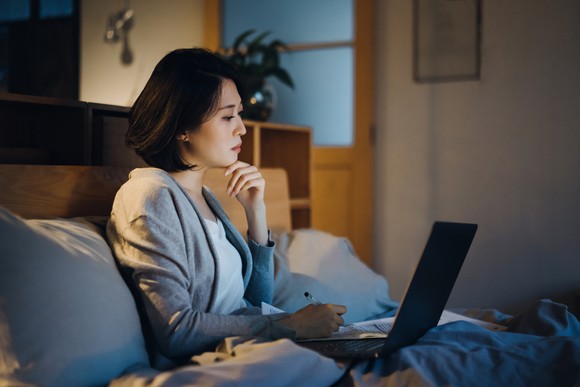 A woman sits in bed while watching a laptop and writing in a notebook.