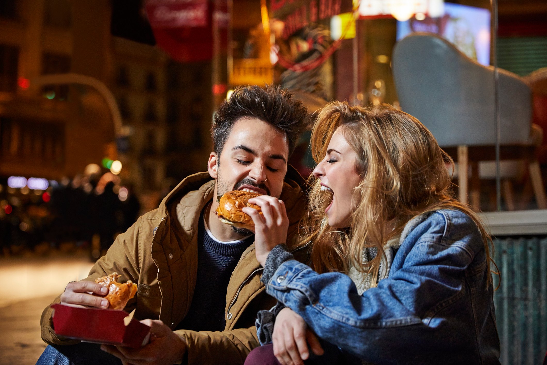 An enthusiasitcally happy woman giving her date a bite out of a hamburger while sitting outside a restaurant in the evening. 
