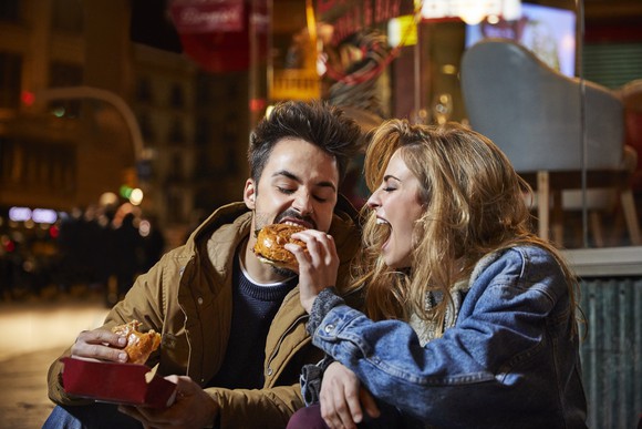 An enthusiasitcally happy woman giving her date a bite out of a hamburger while sitting outside a restaurant in the evening. 