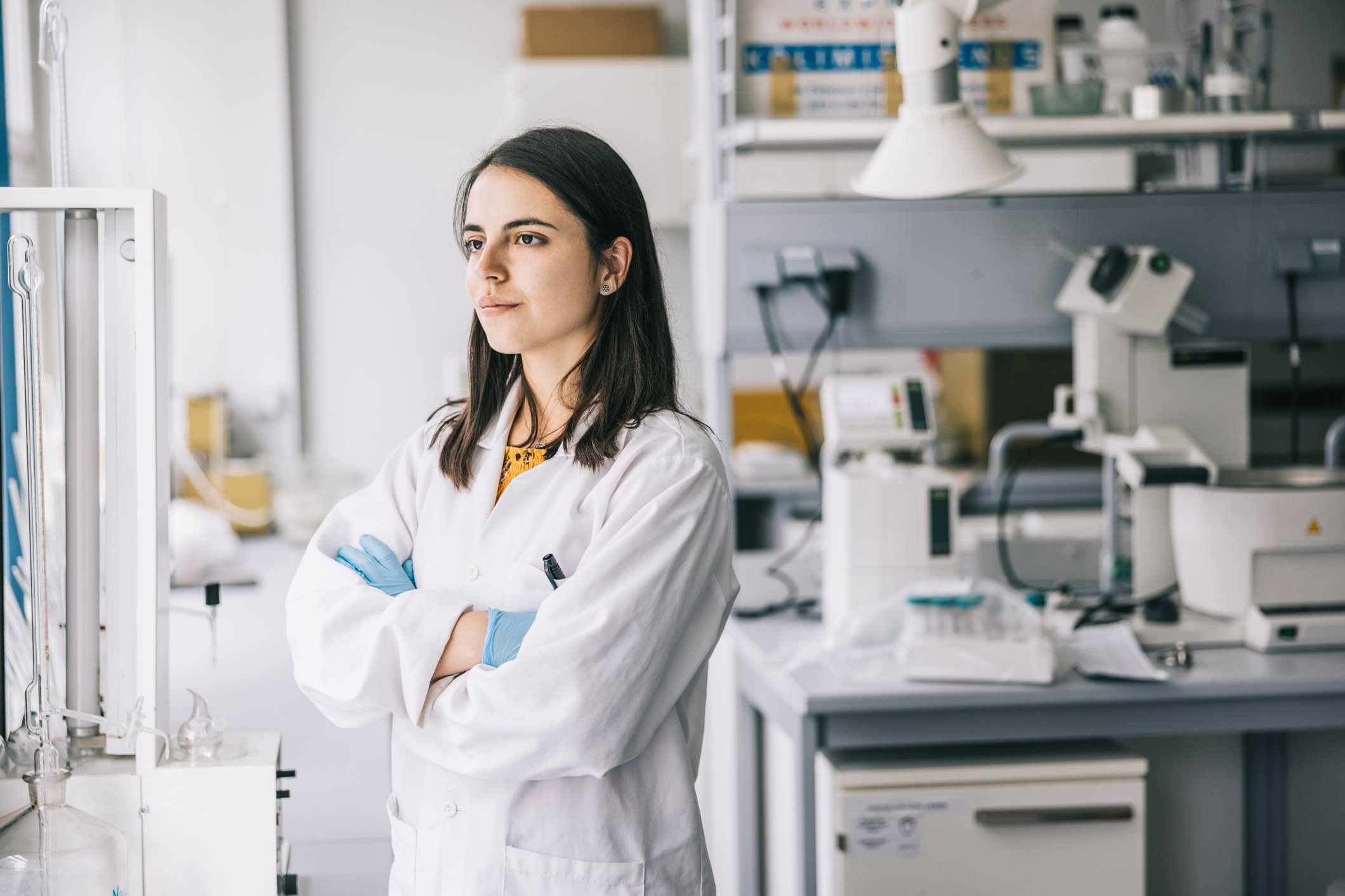 A scientist stands in her laboratory, looking away from her bench while thinking.