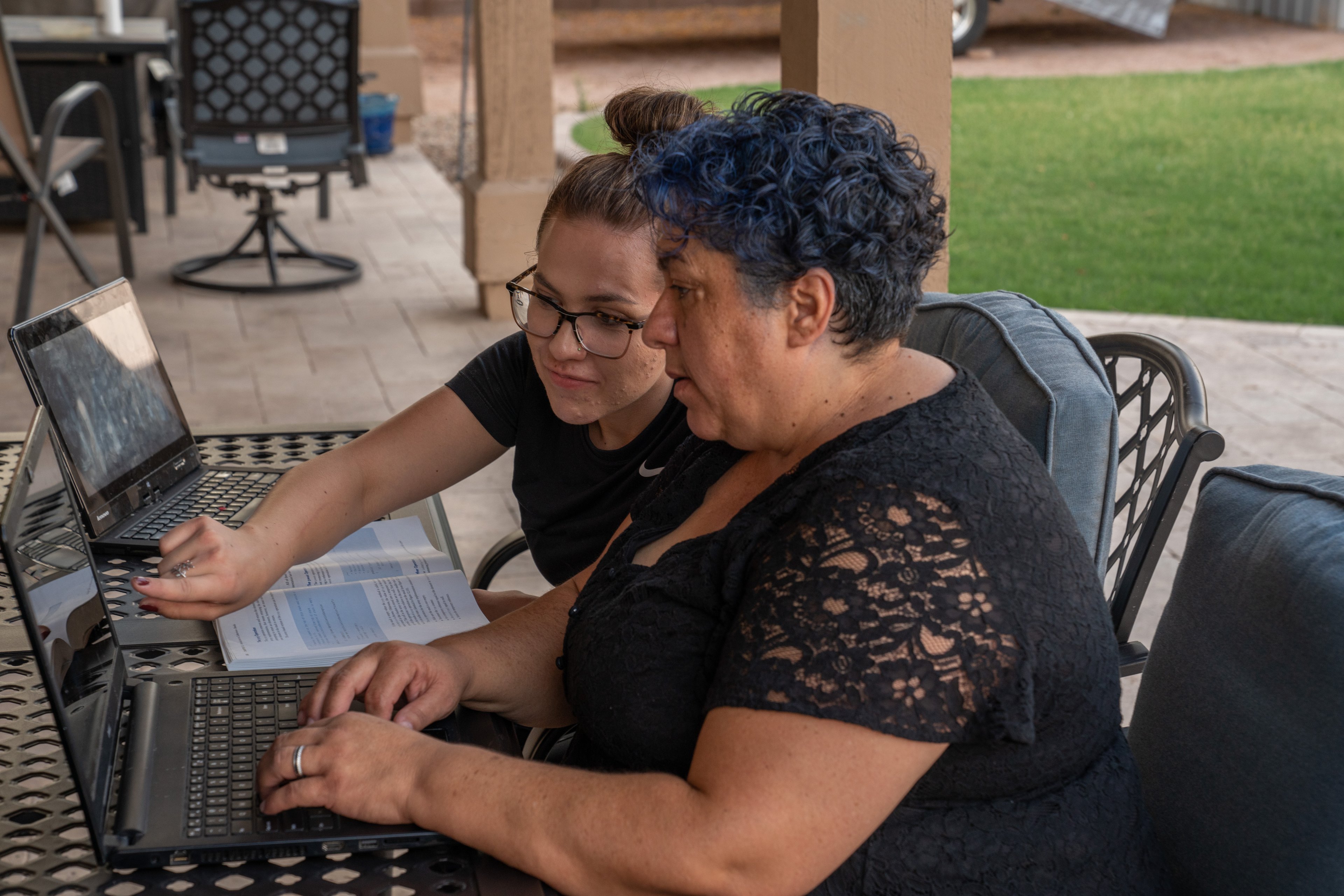 A woman points to a laptop screen while a second woman types on the laptop keyboard. Both women are sitting at a desk outdoors.