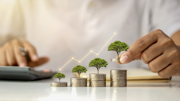 an unidentified person enters figures into a hand calculator sitting on a table while also stacking small trees atop a ascending row of four coin stacks.